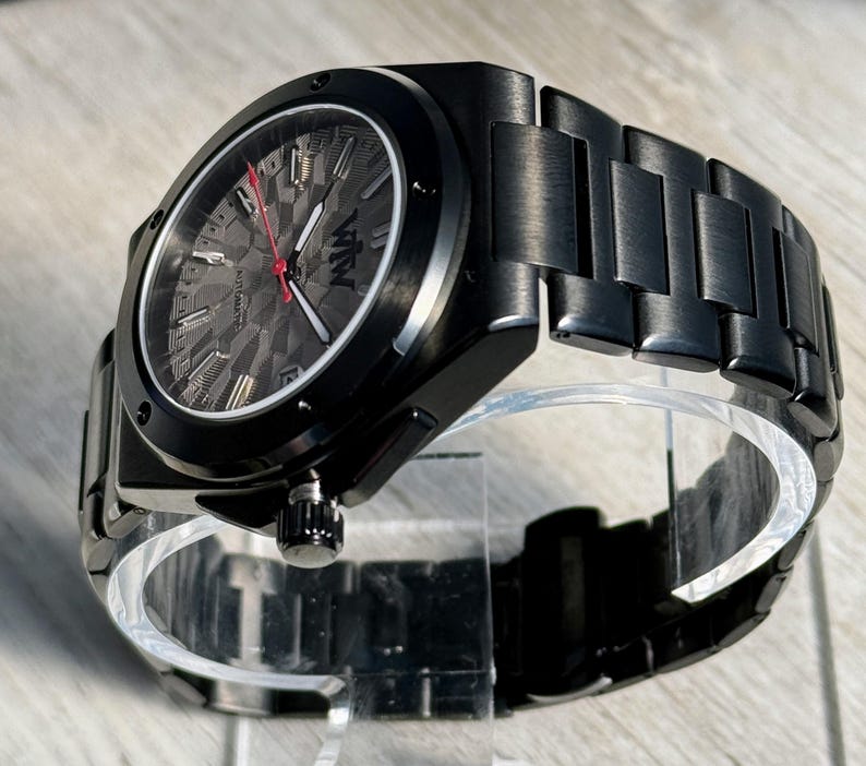K&ouml;nnte beinhalten: Eine schwarze Automatikuhr mit einem matten Metallgliederarmband. Das Zifferblatt hat ein dunkelgraues, strukturiertes Muster, silberfarbene Stundenmarkierungen, wei&szlig;e Zeiger und einen roten Sekundenzeiger. Das Logo VTW ist auf dem Zifferblatt.