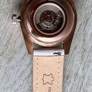 May include: A wristwatch with a rose gold-tone case and a light-coloured leather strap. The back of the watch case is transparent, revealing the internal mechanical movement. The strap is stamped with the text TOP GRAIN LEATHER HAND MADE and 17.