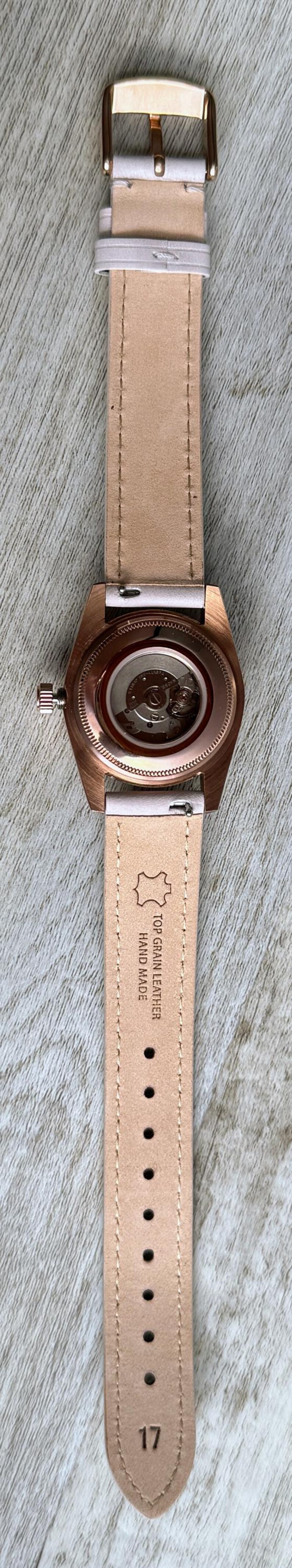 May include: A wristwatch with a rose gold-tone case and a light-coloured leather strap. The back of the watch case is transparent, revealing the internal mechanical movement. The strap is stamped with the text TOP GRAIN LEATHER HAND MADE and 17.