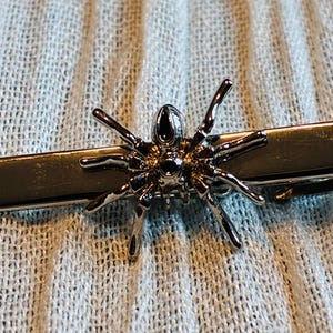 May include: A silver-colored tie clip featuring a detailed spider design. The spider has eight legs and a textured body, attached to a rectangular clip. The clip is designed to secure a tie.