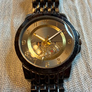 May include: A black wristwatch with a gold-toned face and hands. The watch has a visible mechanical movement and a black metal band. The watch face displays the numbers 1 through 12.