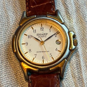 May include: A gold-toned Sharp Quartz wristwatch with a white face, black numerals, and a brown leather band. The watch face displays the time and the text "SHARP QUARTZ" and "WATER RESISTANT".