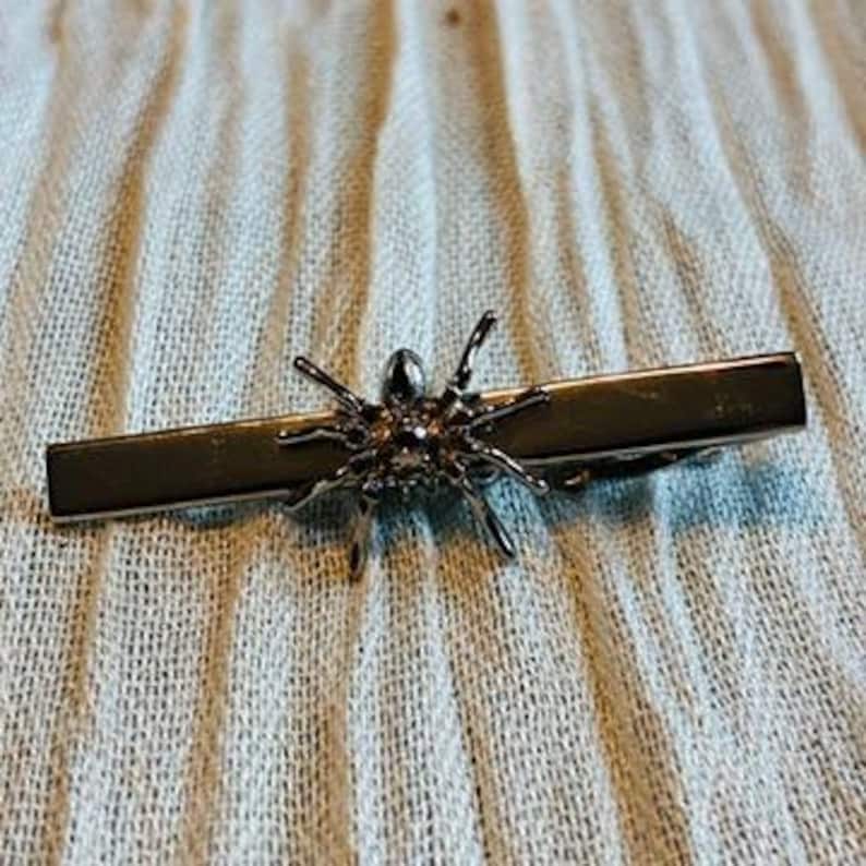 May include: A silver-toned tie bar featuring a detailed spider design. The spider has eight legs and a textured body, attached to a rectangular bar. The tie bar is designed to secure a tie and add a unique accessory.