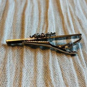 May include: A gold-toned tie clip with a decorative top featuring a series of small, detailed figures. The clip has a hinged design for secure attachment to a tie. The background is a light blue fabric.