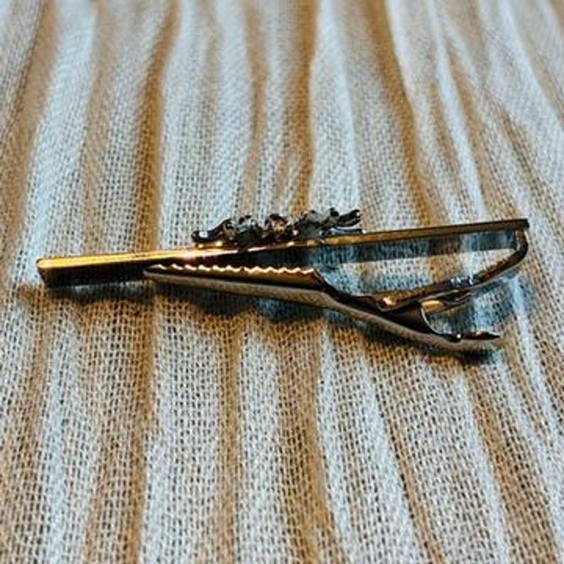 May include: A gold-toned tie clip with a decorative top featuring a series of small, detailed figures. The clip has a hinged design for secure attachment to a tie. The background is a light blue fabric.