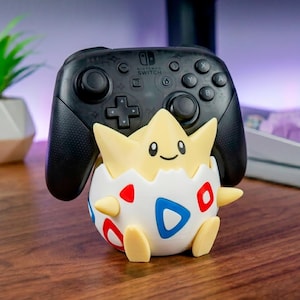 Puede incluir: Un mando Nintendo Switch Pro negro apoyado sobre un soporte con forma de Togepi amarillo y blanco. El soporte muestra una criatura de color amarillo pálido con cara sonriente que emerge de un huevo blanco con patrones triangulares rojos y azules.
