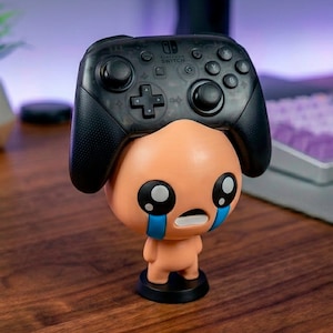 Könnte beinhalten: Eine tanfarbene Figur eines Charakters mit großen schwarzen Augen und blauen Tränen, die einen schwarzen Nintendo Switch Pro Controller hält. Der Controller befindet sich auf dem Kopf der Figur. Die Figur steht auf einem schwarzen Sockel.