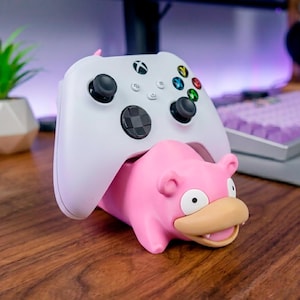 May include: A white wireless video game controller resting on a pink, cartoon-style creature figurine stand. The controller features black analog sticks, a black directional pad, and multicolored action buttons. The pink stand has a tan snout.