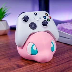 May include: A pink, 3D-printed Mew-shaped controller stand holding a white Xbox controller. The stand has large blue and white eyes. The controller has black joysticks and colorful buttons. The stand is on a wooden surface.