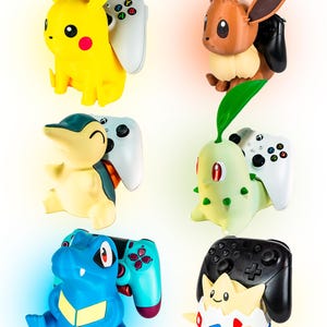 Op de afbeelding: Een verzameling kleurrijke Pokémon-karakterfiguren, elk met een videogamecontroller. De figuren zijn onder meer Pikachu, Eevee, Cyndaquil, Chikorita, Feraligatr en Togepi. De controllers zijn in verschillende kleuren, waaronder wit, zwart en blauwgroen.
