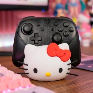 May include: A black Nintendo Switch Pro Controller with a white Hello Kitty-themed charging stand. The stand features a red bow, black whiskers, and a yellow nose. The controller is resting on a wooden surface.