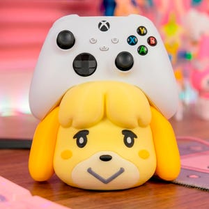 May include: A white Xbox controller rests on top of a yellow figurine of a dog-like character. The controller has black buttons and colored action buttons. The figurine has a smiling face and orange ears.