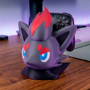 Puede incluir: Un soporte para mando con forma de personaje en gris oscuro y rojo, con cabeza puntiaguda, grandes ojos azules y marcas rojas sobre los ojos y en la parte superior. El soporte está diseñado para sostener un mando de juego negro en su ranura.