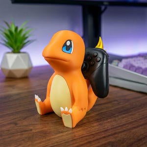 May include: An orange and cream-colored Charmander figurine acting as a controller stand. The character has large blue eyes and white claws on its feet. A black video game controller is held in place by the figure's back and tail area.