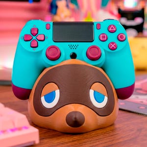 Puede incluir: Un mando de videojuegos turquesa y magenta descansa sobre un soporte marrón y beige con temática de personaje. El mando tiene botones y cruceta magenta, con un panel central negro. El soporte presenta una cara de animal de dibujos animados.