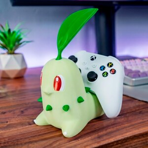 Puede incluir: Un soporte para mando en forma de criatura de color verde claro con una hoja grande y erguida en la cabeza, pequeñas púas en los costados y ojos rojos. Un mando de juego inalámbrico blanco con joysticks negros y botones de colores descansa en él.