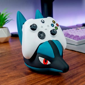 Puede incluir: Un mando Xbox blanco descansa sobre un soporte para mando con temática Lucario en color teal, negro y rojo. El soporte presenta una cabeza de Lucario estilizada con ojos rojos y detalles en teal. El mando tiene botones negros.