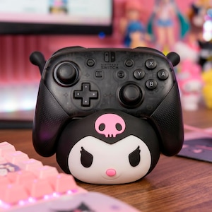 Może przedstawiać: Czarny kontroler Nintendo Switch Pro z podstawką ładującą w stylu Kuromi. Kontroler ma czarne wykończenie z logo Nintendo Switch i układem przycisków. Podstawka ma czarno-biały wzór postaci Kuromi z różową czaszką.