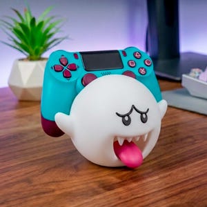 Puede incluir: Un soporte para mando en forma de fantasma blanco y esférico, con la boca abierta, dientes afilados y una lengua rosa que sobresale. Un mando de videojuego de color turquesa y morado oscuro descansa sobre la cabeza de la figura del fantasma.