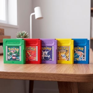 Può includere: Cinque cartucce Game Boy colorate sono esposte su un tavolo di legno. Ogni cartuccia presenta un colore diverso: verde, rosso, viola, giallo e blu. Il logo Pokémon è visibile su ogni cartuccia.