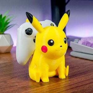 Peut inclure: Une figurine jaune de Pikachu conçue comme support de manette, avec des oreilles aux pointes noires, des joues rouges circulaires et des yeux noirs. Une manette de jeu vidéo blanche est placée derrière le personnage, appuyée contre son dos.