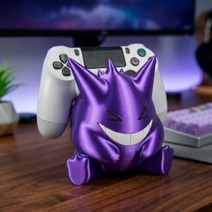 May include: A purple 3D-printed Gengar figure with a white PlayStation controller in the background. The Gengar figure has a smiling face with sharp teeth and pointed ears. The controller is white with black buttons.
