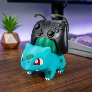 May include: A teal and green creature-shaped controller stand featuring a bulbous plant-like structure on its back. A black wireless game controller with dual analog sticks, a directional pad, and four action buttons is resting securely in the stand.