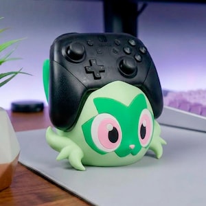 Könnte beinhalten: Ein schwarzer Nintendo Switch Pro Controller auf einem hellgrünen, charakterförmigen Ständer. Der Ständer hat große rosa-weiße Augen, eine kleine Nase und stilisierte blattartige Anhängsel an den Seiten, die an eine fiktive Kreatur erinnern.