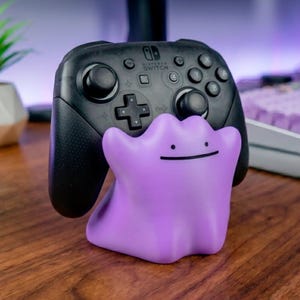 Könnte beinhalten: Ein schwarzer Nintendo Switch Pro Controller auf einem violetten, klecksförmigen Charakter-Ständer. Der Ständer hat ein einfaches Gesicht mit zwei kleinen schwarzen Punkten als Augen und einem dünnen, horizontalen schwarzen Strich als Mund.