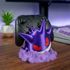 May include: A purple Gengar-shaped controller stand featuring red eyes and a white grin, resting on a light purple cloud-like base. A black Nintendo Switch Pro controller is docked securely within the top of the character figure.