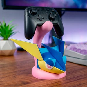 Könnte beinhalten: Ein rosa-blauer Videospiel-Controller-Ständer mit einem stilisierten Design. Der Ständer hält einen schwarzen Controller. Das Design umfasst gelbe und blaue Akzente und steht auf einer Holzoberfläche.