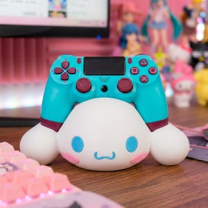 May include: A teal and burgundy PlayStation 4 controller rests on top of a white, cartoon-style character with pink cheeks and blue accents. The controller has a black touchpad and the character has a smiling face. A pink keyboard is in the foreground.