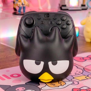 Puede incluir: Un soporte para mando negro con la forma del personaje Badtz-Maru, con grandes ojos blancos y un pico amarillo, que sostiene un mando negro Nintendo Switch Pro con botones estándar, joysticks y el logotipo de Nintendo Switch en el centro.