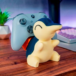 Könnte beinhalten: Ein hellblauer Gaming-Controller ruht auf einem cremefarbenen und dunkelblauen, charakterförmigen Ständer. Der Ständer zeigt eine stilisierte Kreatur mit glatter, matter Oberfläche, einem runden Kopf und einer spitzen Schnauze zur Halterung.