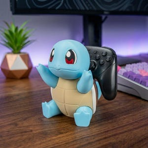 May include: A light blue and tan Squirtle character figure designed as a controller stand. The figure has large red eyes and is holding a black gaming controller in its arms. The character is seated in an upright position with its arms extended.