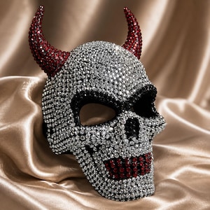 Könnte beinhalten: Eine Totenkopfmaske, die mit klaren und roten Strasssteinen besetzt ist. Die Maske hat zwei markante rote Strasshörner an der Oberseite sowie schwarze Strassakzente um die Augenhöhlen und rote Strasssteine, die die Zähne und den Kiefer bilden.