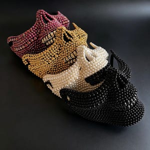 May include: A collection of four skull-shaped masks, each covered in small, spherical beads. The masks are arranged in a row, featuring distinct colors: a deep red, a metallic gold, a creamy white, and a solid black, all with defined teeth.