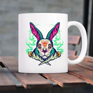 Może przedstawiać: Biały ceramiczny kubek z kolorową ilustracją czaszki królika. Projekt zawiera odcienie turkusu, różu i pomarańczy, z zielonymi wzorami przypominającymi dym. Królik ma serce na czole. Kubek ma biały uchwyt.