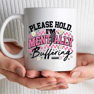 May include: White ceramic mug with a large handle, featuring the text "PLEASE HOLD I'M MENTALLY BUFFERING" in black and pink. The design includes a lightning bolt and daisy accents.