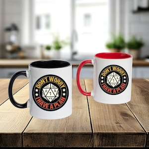 Puede incluir: Dos tazas de café blancas con asas negras y rojas. Cada taza presenta el texto "DON'T WORRY I HAVE A PLAN" en un diseño circular con un gráfico de dados. El interior de una taza es negro, la otra es roja.