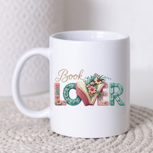 May include: White ceramic mug featuring the text "Book Lover" in a decorative font. The "O" in "Book" is a book with flowers. The letters are in a knitted style with pink, teal, and gold accents. A lovely gift for book enthusiasts.