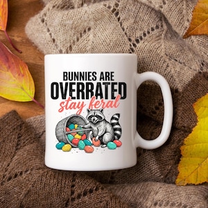 Puede incluir: Una taza de cerámica blanca con asa negra y el texto "But First, Coffee" en negrita negra. La taza se encuentra sobre un fondo borroso, sugiriendo un ambiente acogedor.