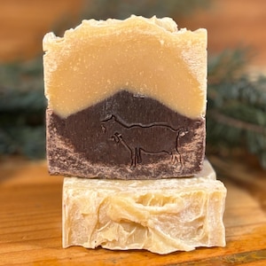 May include: Two-tone goat milk soap bars. The top layer is a light tan color, and the bottom layer is a dark brown with a goat silhouette. The soap bars are stacked on top of each other. The background includes a pine cone and evergreen branches.