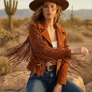 May include: A brown suede jacket featuring long fringe detailing on the sleeves, worn with a brown felt hat, a white tank top, blue denim jeans, and a brown leather belt with a large silver-toned buckle, captured in a desert setting.