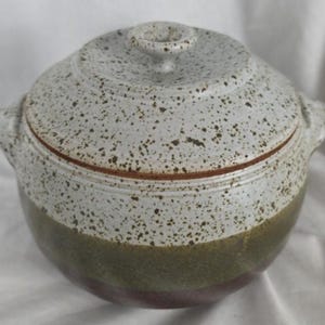 May include: A speckled ceramic lidded pot with two handles. The pot has a white and green exterior with brown speckles. The lid has a small handle on top. The bottom of the pot is a dark red color.