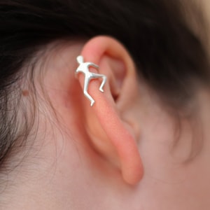 以下が含まれることがあります： 人が登っている形のシルバーのイヤーカフ。カフは耳の上部に装着されています。