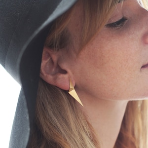 Könnte beinhalten: Eine Frau trägt einen schwarzen Hut mit einem goldenen geometrischen Ohrring. Der Ohrring hat eine scharfe, dreieckige Form.