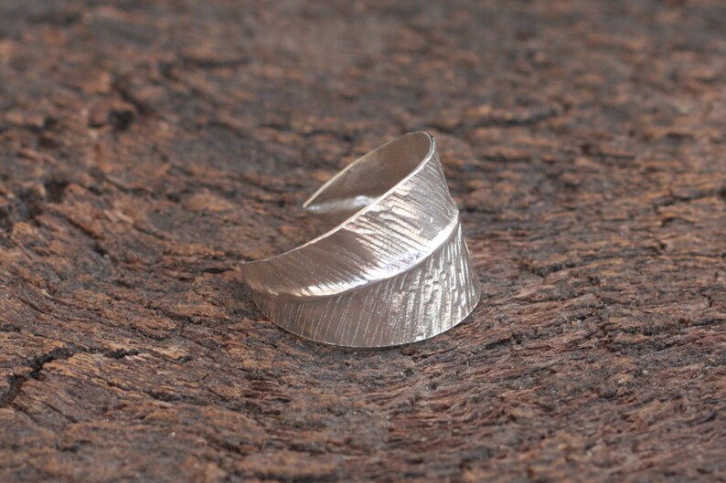 May include: A silver ring with a textured, leaf-shaped design. The ring is open and adjustable.