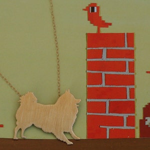 Op de afbeelding: Een gouden ketting met een silhouet van een hond die eraan hangt. De ketting is tegen een groene achtergrond met een rode bakstenen muur en rode vogels bovenop de muur.