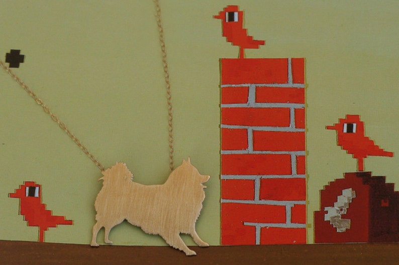 Op de afbeelding: Een gouden ketting met een silhouet van een hond die eraan hangt. De ketting is tegen een groene achtergrond met een rode bakstenen muur en rode vogels bovenop de muur.